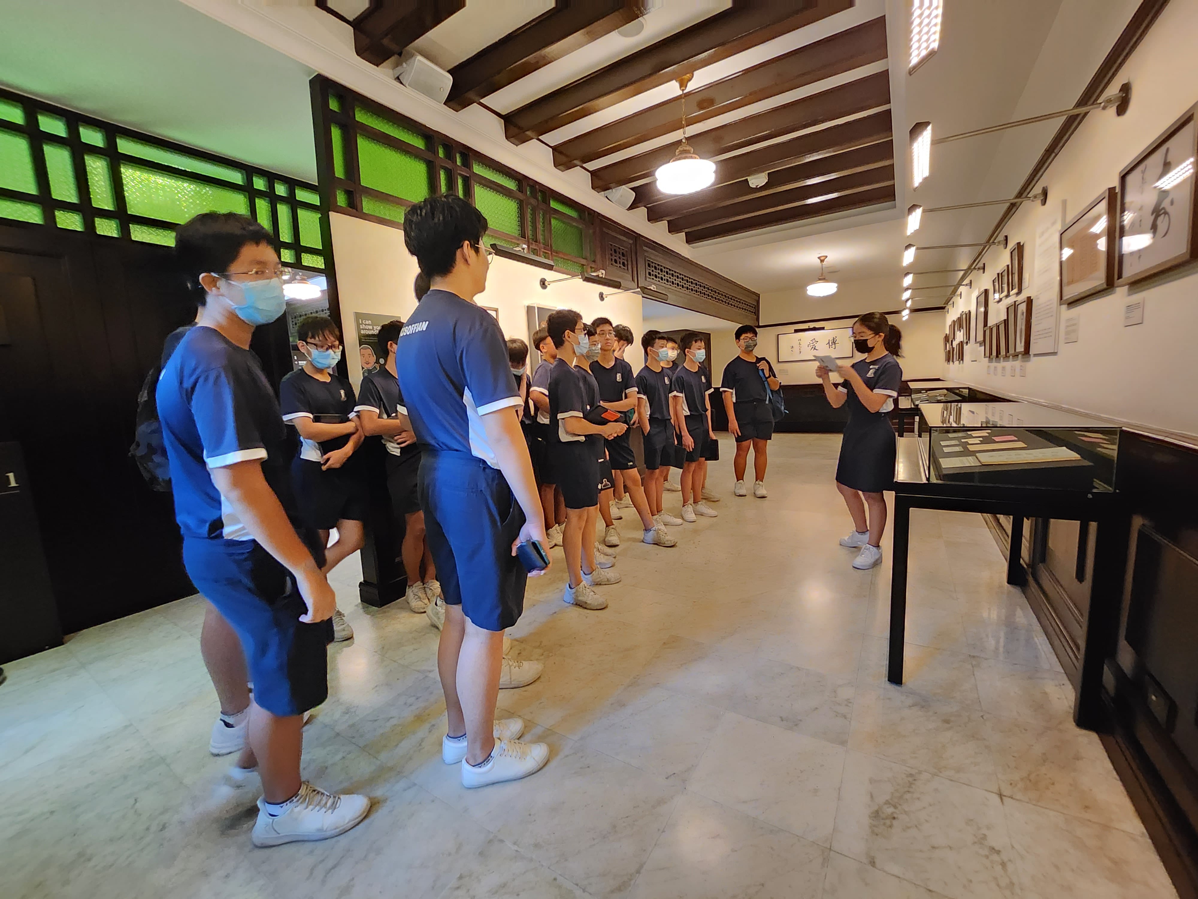 Students in blue uniforms and face masks stand in a museum hall listening to a guide near display cases.